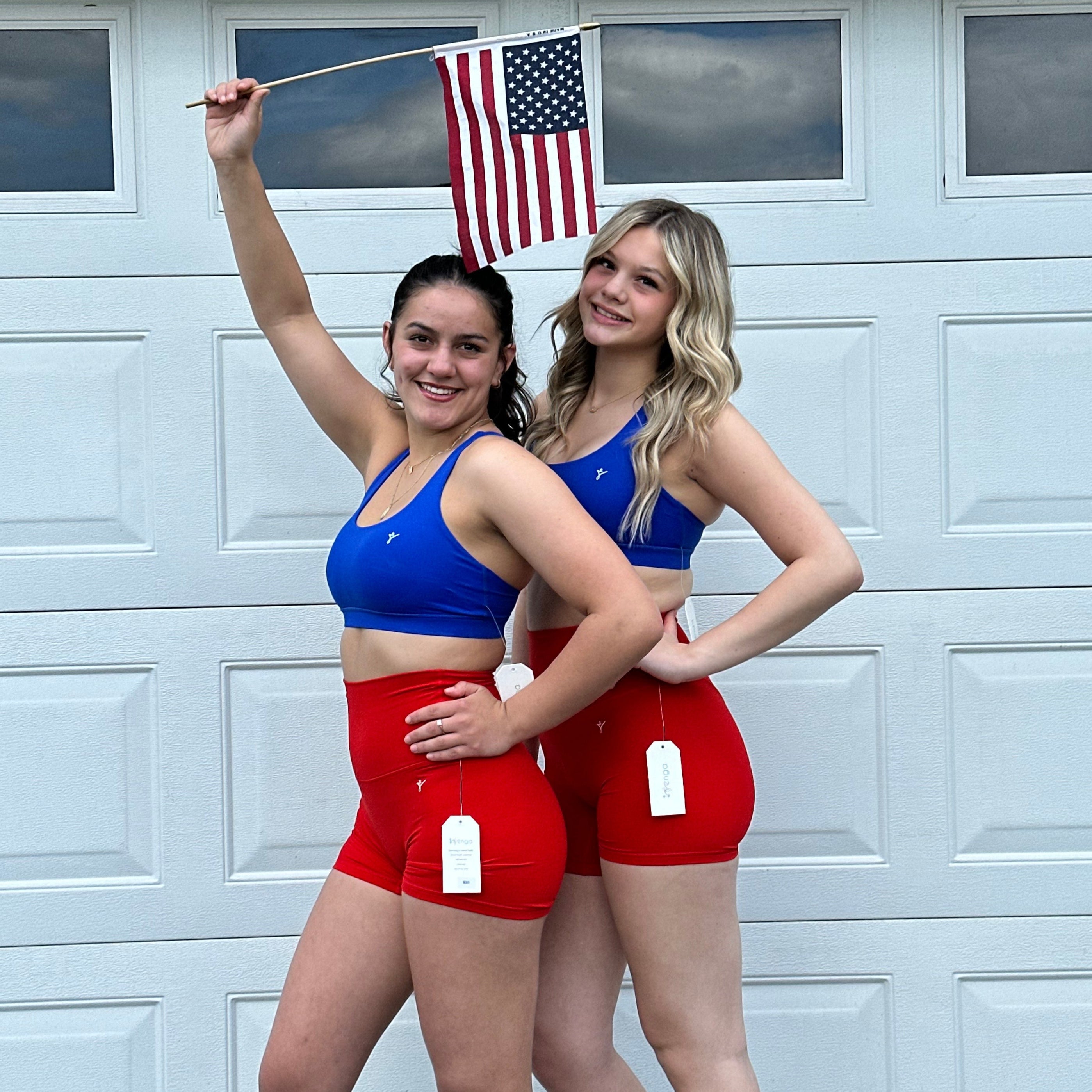 Two women in matching blue sports bras and red shorts holding an American flag in front of a garage.